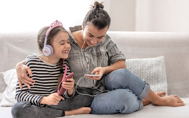 Madre e hija alegres descansan en casa, escuchando música en auriculares