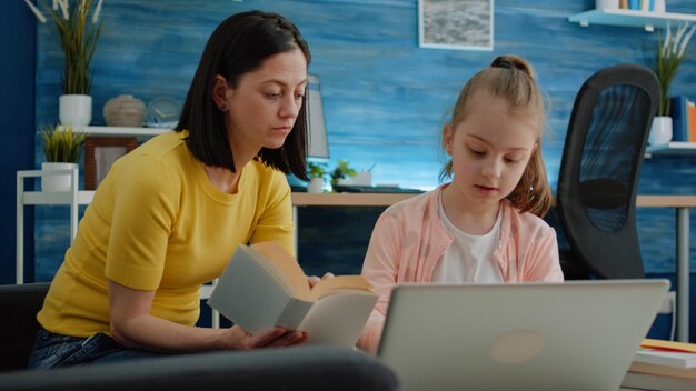 Madre e hija aprendiendo junto con libros y computadora portátil para educación remota. Niño que usa el dispositivo para estudiar cursos y lecciones en línea. Niña de la escuela primaria estudiando con sus padres