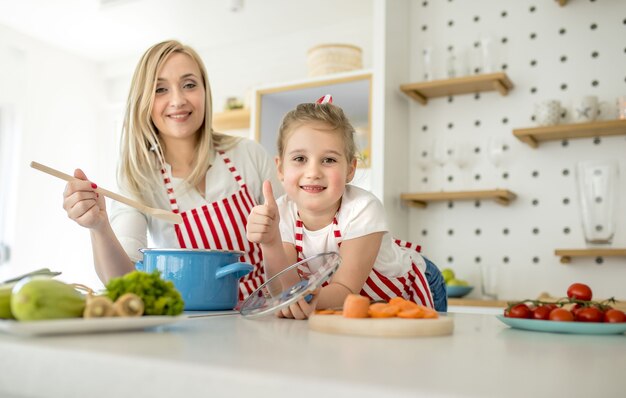 Madre e hija caucásicas vistiendo delantales a juego sonriendo y posando en una cocina