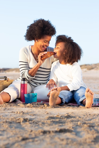 Madre e hija divirtiéndose en la playa. Madre e hija con ropa informal sentadas en una manta, comiendo, mujer alimentando a un niño. Familia, relajación, concepto de naturaleza.