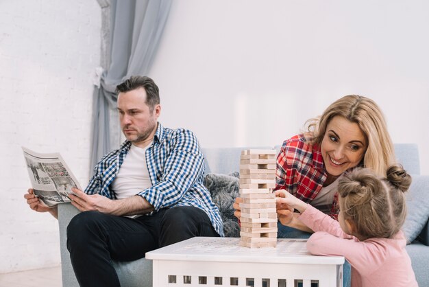 Madre e hija felices que juegan al juego de madera del bloque mientras que padre lee el periódico en casa