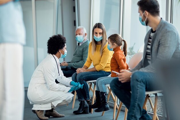 Madre e hija hablando con un médico afroamericano mientras usan máscaras faciales en la sala de espera del hospital