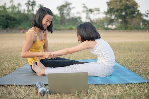 Madre e hija haciendo yoga. Entrenamiento de mujeres y niños en el parque. Deportes al aire libre. estilo de vida deportivo saludable, ver el video tutorial en línea de ejercicios de yoga y pose de flexión hacia adelante sentado.