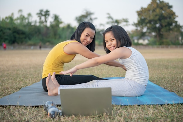 Madre e hija haciendo yoga. Entrenamiento de mujeres y niños en el parque. Deportes al aire libre. estilo de vida deportivo saludable, ver el video tutorial en línea de ejercicios de yoga y pose de flexión hacia adelante sentado.