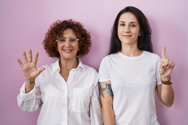 Foto gratuita madre e hija hispanas con pantalones blanco informal sobre fondo rosa que aparecen y señalan con los dedos número seis mientras sonríen confiadas y felices.