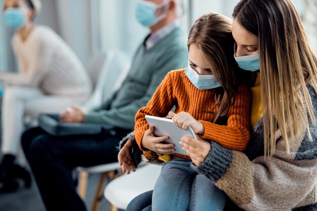 Madre e hija con mascarillas usando el panel táctil mientras esperan en la clínica médica