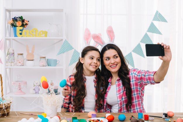 Madre e hija con orejas de conejo tomando selfie en teléfono móvil en casa