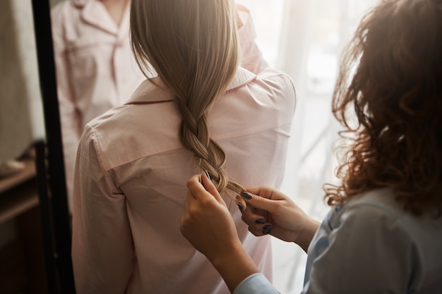 Madre e hija de pie en ropa de dormir mientras mamá haciendo trenzas para su chica, peinándose y mirándose en el espejo