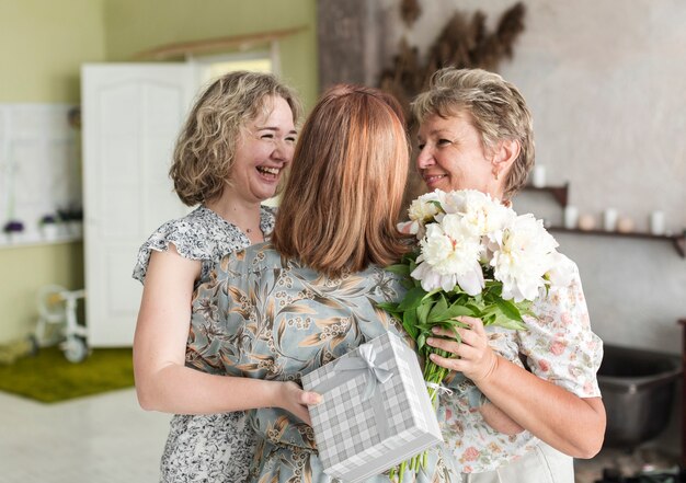 Madre e hija que abrazan a su abuela con sostener el ramo del regalo y de la flor en casa