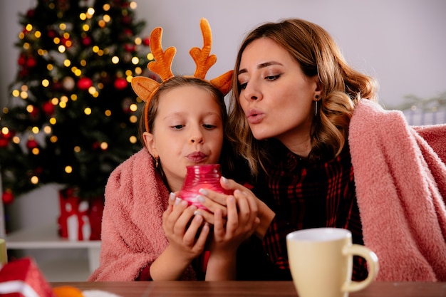 Foto gratuita madre e hija satisfechas con manta sosteniendo y soplando velas sentado en la mesa disfrutando de la época navideña en casa