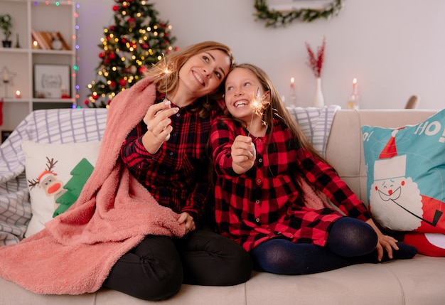 Madre e hija satisfechas sosteniendo bengalas cubiertas con una manta sentadas en el sofá y disfrutando de la Navidad en casa