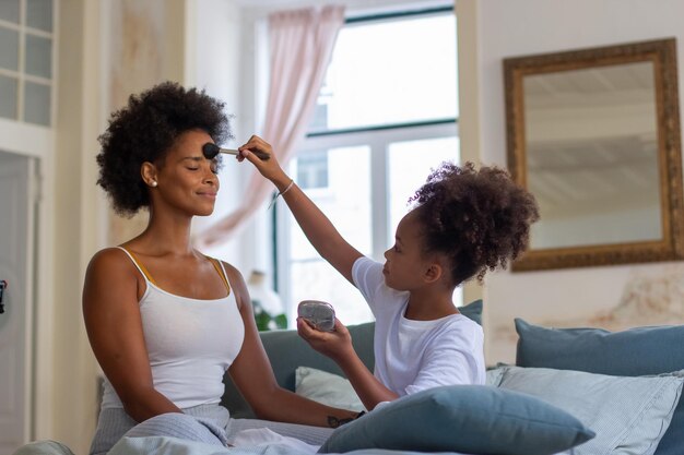 Madre e hija sonrientes maquillándose juntas. Niña enfocada con el pelo rizado sosteniendo un cepillo, aplicando maquillaje en la frente de mamá. Familia, unión, ocio, concepto de hobby.