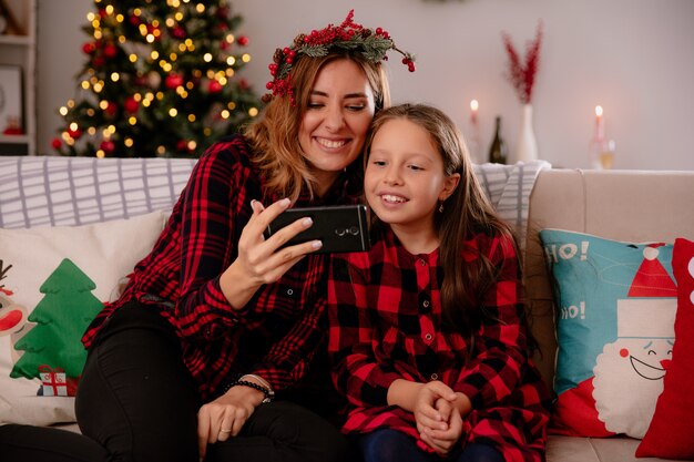 Madre e hija sonrientes mirando el teléfono sentados en el sofá y disfrutando de la Navidad en casa