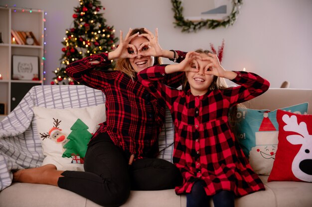 Madre e hija sonrientes mirando a través de los dedos sentados en el sofá y disfrutando de la Navidad en casa