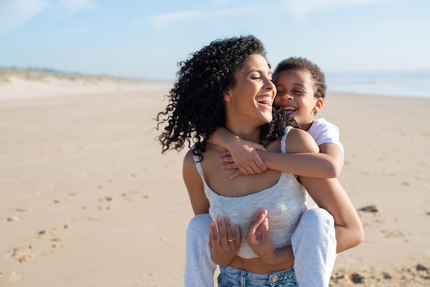 Madre e hijo alegres pasando tiempo en la playa. Familia afroamericana caminando, riendo, jugando, montando en la espalda. Ocio, tiempo en familia, concepto de paternidad.