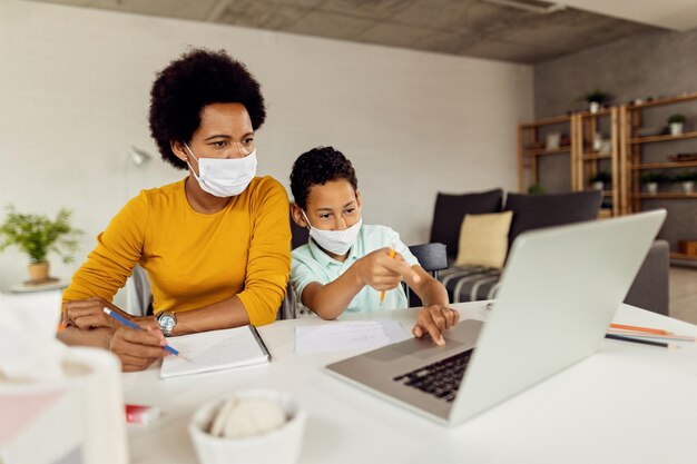 Madre e hijo negros con máscaras faciales usando una computadora portátil mientras estudian en casa