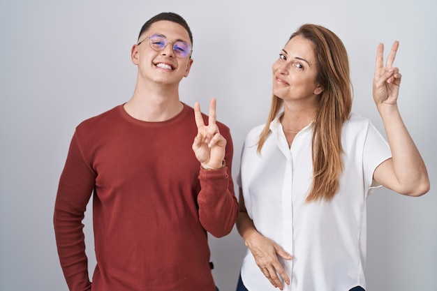 Madre e hijo parados juntos sobre un fondo aislado sonriendo mirando a la cámara mostrando los dedos haciendo el signo de la victoria. número dos.