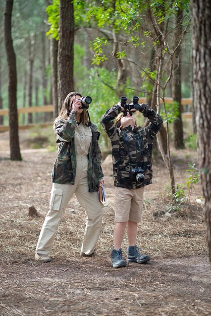 Madre e hijo preparándose para tomar fotografías en el bosque. Familia con cámaras modernas usando binoculares, de pie. Crianza, familia, concepto de ocio.