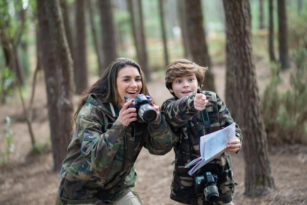 Madre e hijo sorprendidos tomando fotos en el bosque. Familia con cámaras modernas apuntando, mirando con los ojos bien abiertos. Crianza, familia, concepto de ocio.