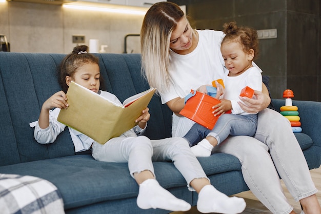 Madre e hijos relajándose juntos en el sofá de casa en la sala de estar. Niñas leyendo un libro y jugando juguetes.