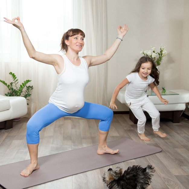 Madre embarazada haciendo entrenamiento de yoga de diversión con su niña de cabrito y mascota jugando en casa
