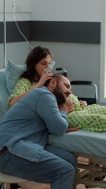 Madre embarazada tendido en la cama de la sala de hospital con equipo médico y marido sosteniendo la mano sobre el vientre. Enfermera afroamericana que controla la atención médica durante el embarazo para asistencia en el parto