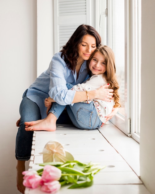 Foto gratuita madre feliz e hija que abrazan en travesaño de la ventana