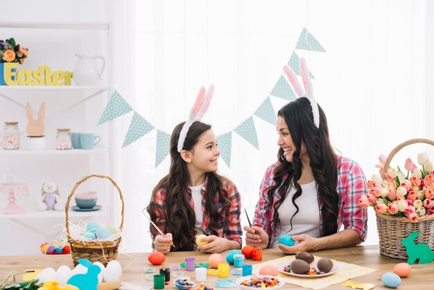 Madre feliz e hija que pintan los huevos de Pascua con el cepillo en casa