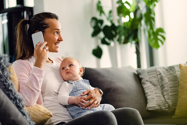 Madre feliz hablando por teléfono mientras sostiene a su bebé y se relaja en el sofá de la sala de estar