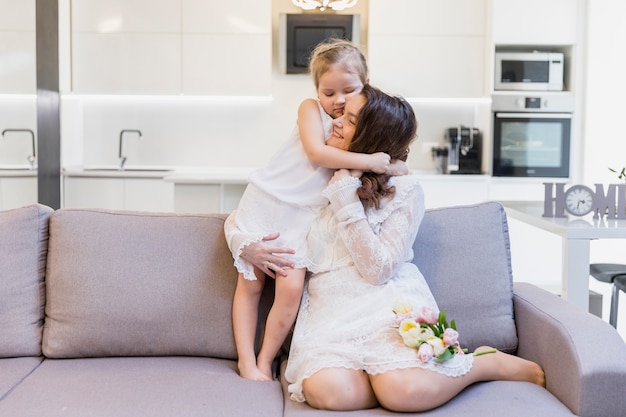 Madre feliz que abraza a su niña linda en el sofá en sala de estar