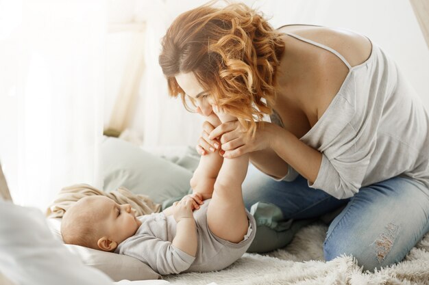 Madre feliz que juega con el bebé recién nacido que besa las patitas que pasan los mejores momentos de maternidad en el dormitorio acogedor. Cálido ambiente familiar. Concepto de familia