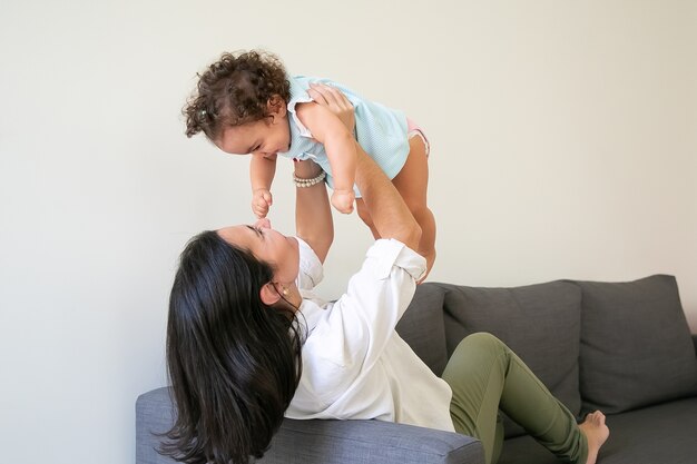 Madre feliz sosteniendo a la niña en brazos, levantando al niño mientras está sentado en el sofá en casa. Concepto de paternidad e infancia