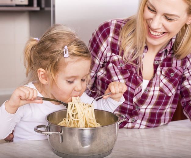 Madre feliz con su hija comiendo espaguetis caseros en la encimera de la cocina