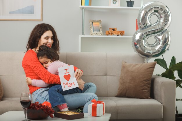 Madre feliz con su pequeña hija sentada en un sofá sosteniendo una tarjeta de felicitación sonriendo alegremente en la sala de estar iluminada celebrando el día internacional de la mujer el 8 de marzo