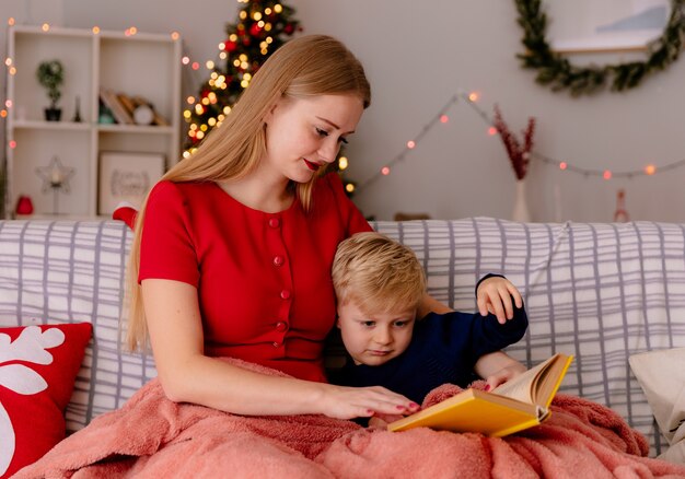 Madre feliz en vestido rojo con su pequeño hijo debajo del libro de lectura de la manta en una habitación decorada con árbol de Navidad en la pared