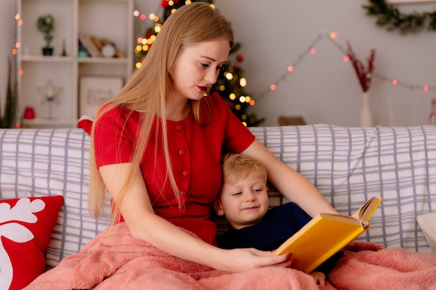 Madre feliz en vestido rojo con su pequeño hijo debajo de una manta con un libro leyendo en una habitación decorada con árbol de Navidad en la pared