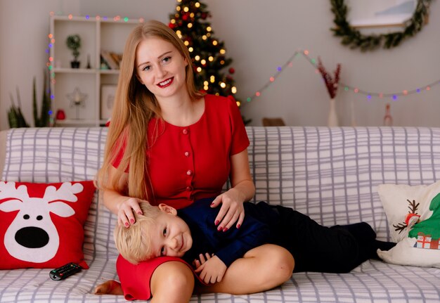 Madre feliz en vestido rojo con su pequeño hijo que está acostado de rodillas en un sofá divirtiéndose viendo la televisión juntos en una habitación decorada con árbol de Navidad en el fondo