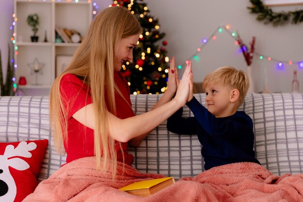 Madre feliz en vestido rojo con su pequeño niño debajo de la manta con un libro divirtiéndose dando cinco en una habitación decorada con árbol de Navidad en la pared