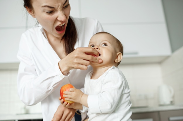 Madre hermosa joven que alimenta con melocotón a su pequeño hijo en la cocina. Mamá haciendo una expresión divertida para hacer reír a su hijo.