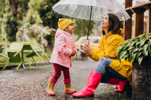 Madre con hija caminando en el parque bajo la lluvia con botas de goma