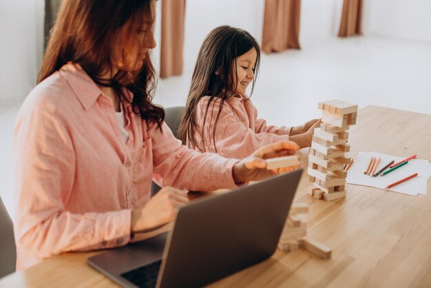 Madre con hija jugando jenga en casa y usando laptop