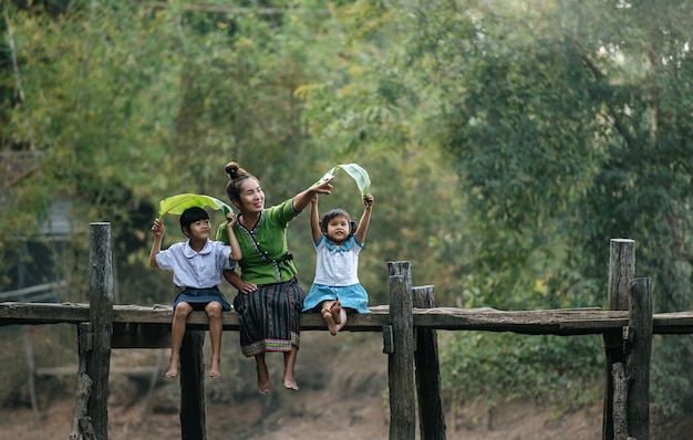 Madre joven asiática sentada en un puente de madera y disfruta jugando con dos hijas pequeñas en uniforme de estudiante con hoja de plátano, espacio para copiar, concepto de estilo de vida rural
