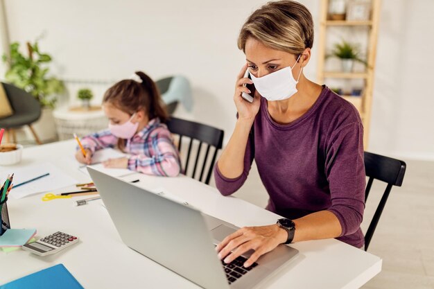 Madre joven con cara protectora hablando por teléfono mientras trabaja en una laptop en casa