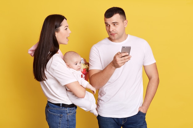 Madre joven enojada que sostiene al bebé en las manos y que mira a su marido usando el teléfono