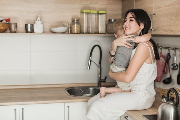 Madre joven feliz que lleva a su hijo que se sienta en contador de la cocina