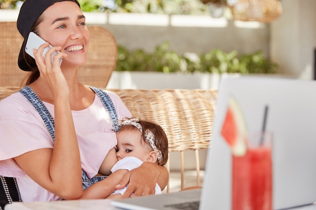 Una madre joven positiva de ojos azules le da leche materna a su pequeño bebé, habla con alguien a través del teléfono celular y da consejos sobre cómo cuidar a los niños