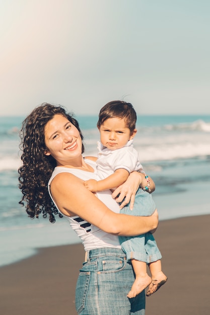 Madre joven que celebra al bebé en manos en la playa