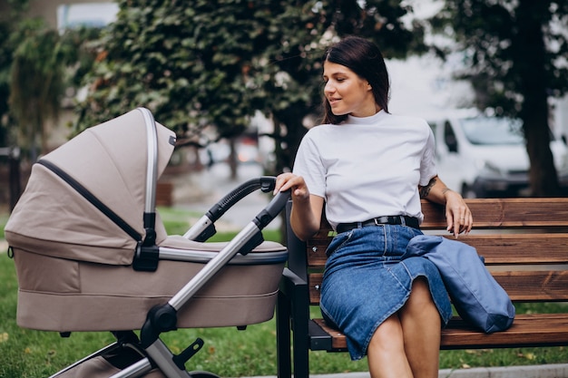 Madre joven sentada en un banco en el parque con cochecito de bebé