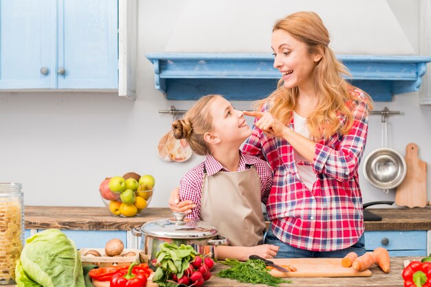 Madre joven sonriente que toca la nariz de su hija con el dedo en la cocina