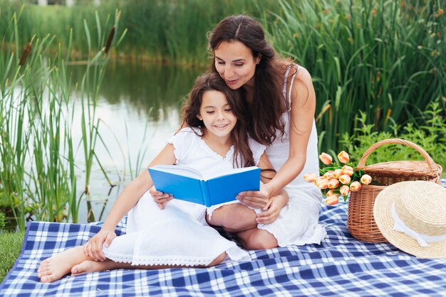 Madre leyendo libro a su hija junto al lago.